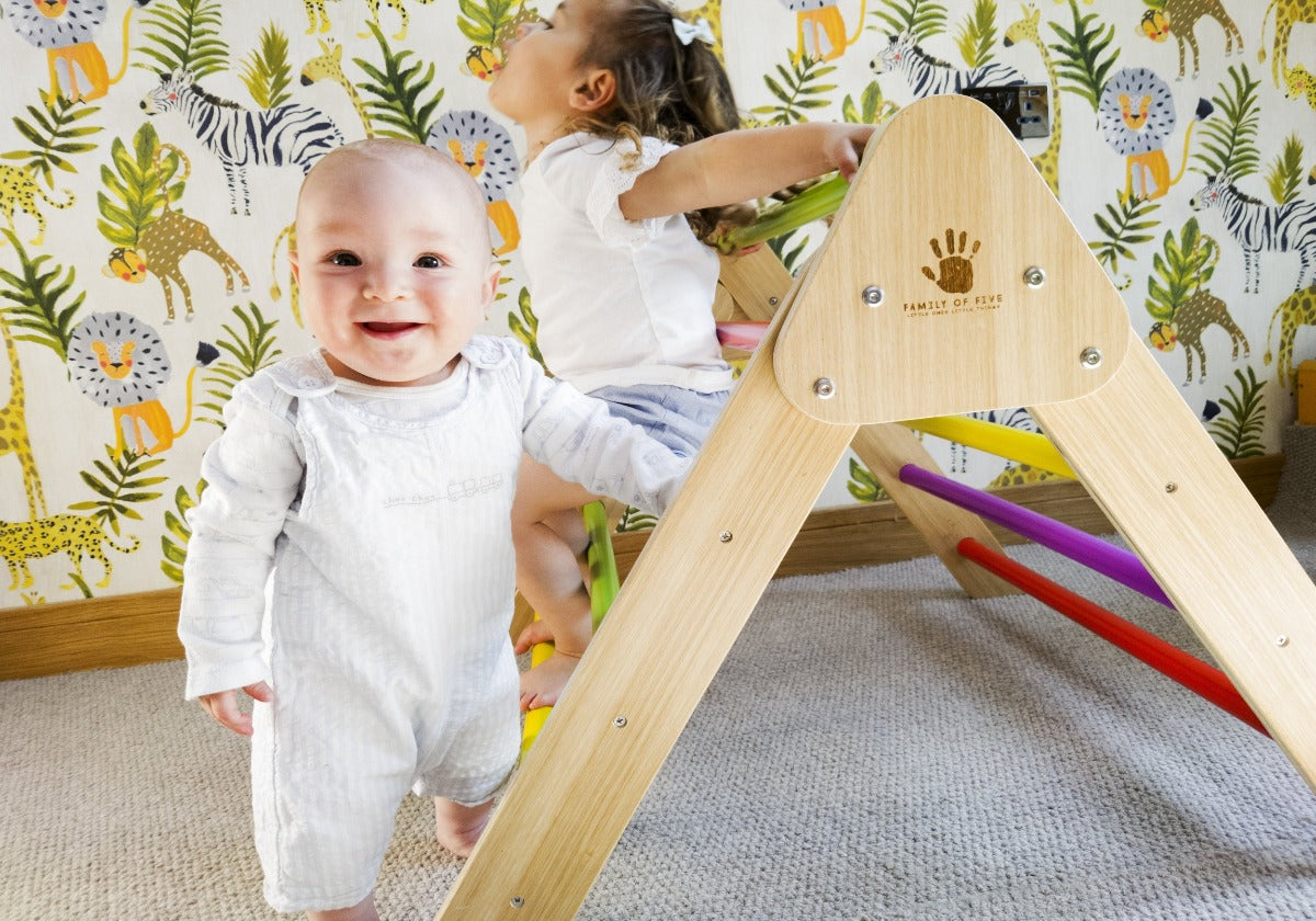 Little boy using the side of a pikler climbing frame to support himself standing up whilst a little girl sits on the toddler climbing frame bars