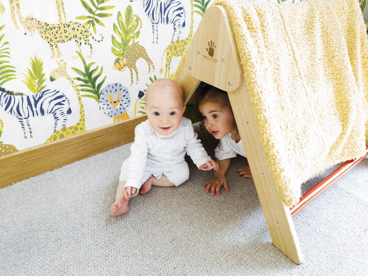 Little boy sitting on the floor infront of a toddler climbing frame. The  frame has a blanket over it to look like a tent and there is a little girl inside